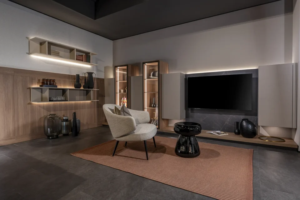 Modernes Wohnzimmer mit dunklem Boden, grauer Wand und Holzakzenten. Ein beiger Sessel steht auf einem roten Teppich vor einem schwarzen Couchtisch. An der Wand ein großer Fernseher und beleuchtete Regale mit Dekorationsobjekten.