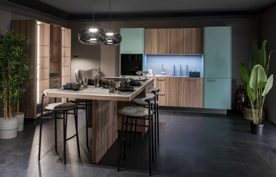 Moderne Küche mit Holz- und türkisfarbenen Schränken. In der Mitte steht eine Insel mit vier Hockern. Zwei schwarze Hängeleuchten sind über der Insel. Links eine große Pflanze, rechts ein hoher Pflanzentopf. Beleuchtung vom Küchenrückwandbereich.