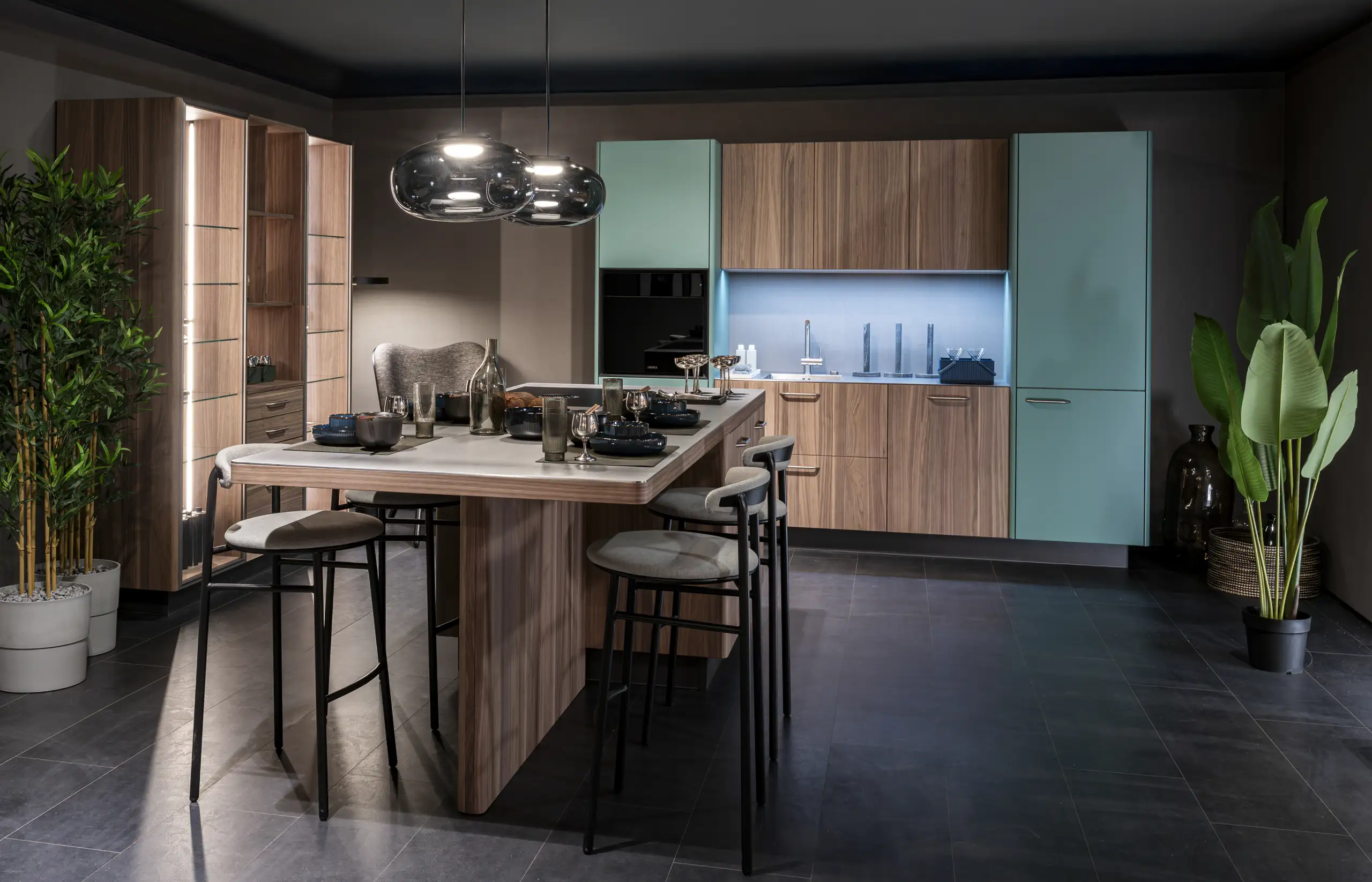 Moderne Küche mit Holz- und türkisfarbenen Schränken. In der Mitte steht eine Insel mit vier Hockern. Zwei schwarze Hängeleuchten sind über der Insel. Links eine große Pflanze, rechts ein hoher Pflanzentopf. Beleuchtung vom Küchenrückwandbereich.