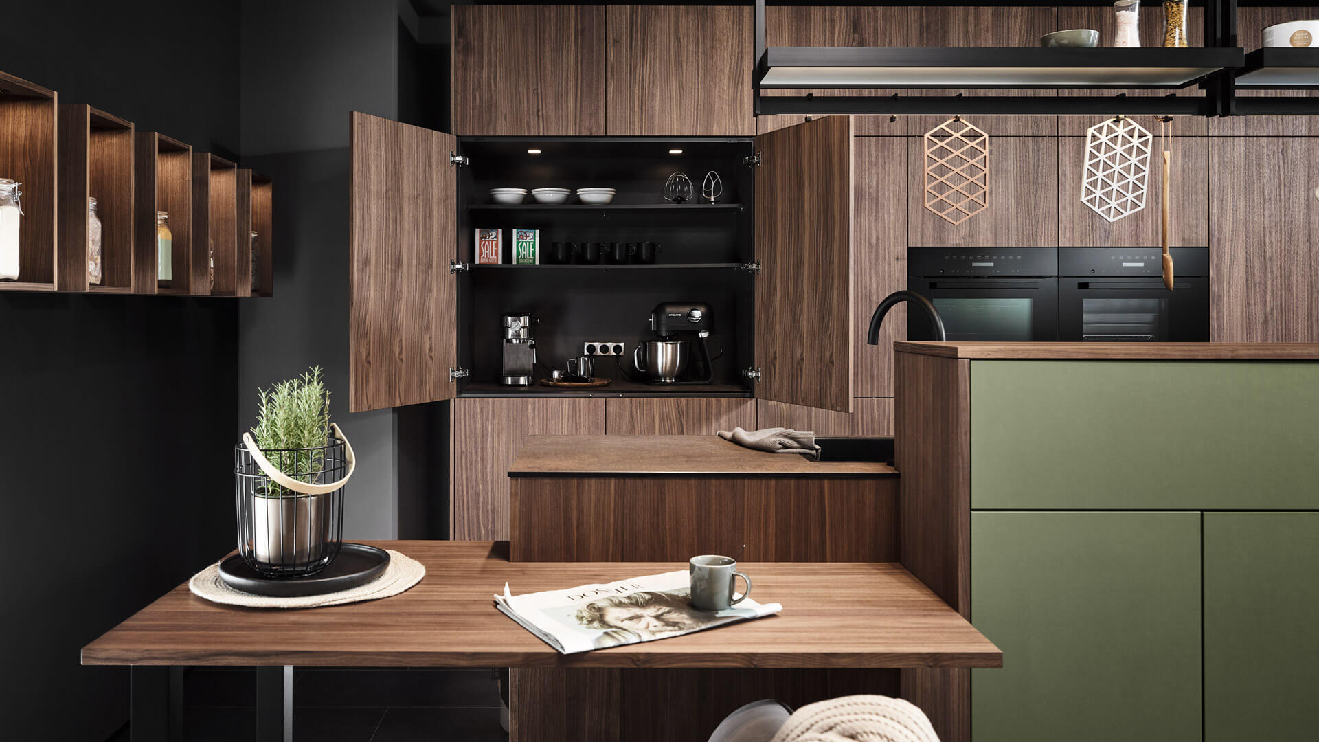 Moderne Küche mit dunklen Holzschränken, offenem Regal, integriertem Backofen und Kaffeemaschine. Ein Tisch mit Pflanze, Zeitschrift und Tasse steht im Vordergrund. Dezente Beleuchtung sorgt für eine gemütliche Atmosphäre.