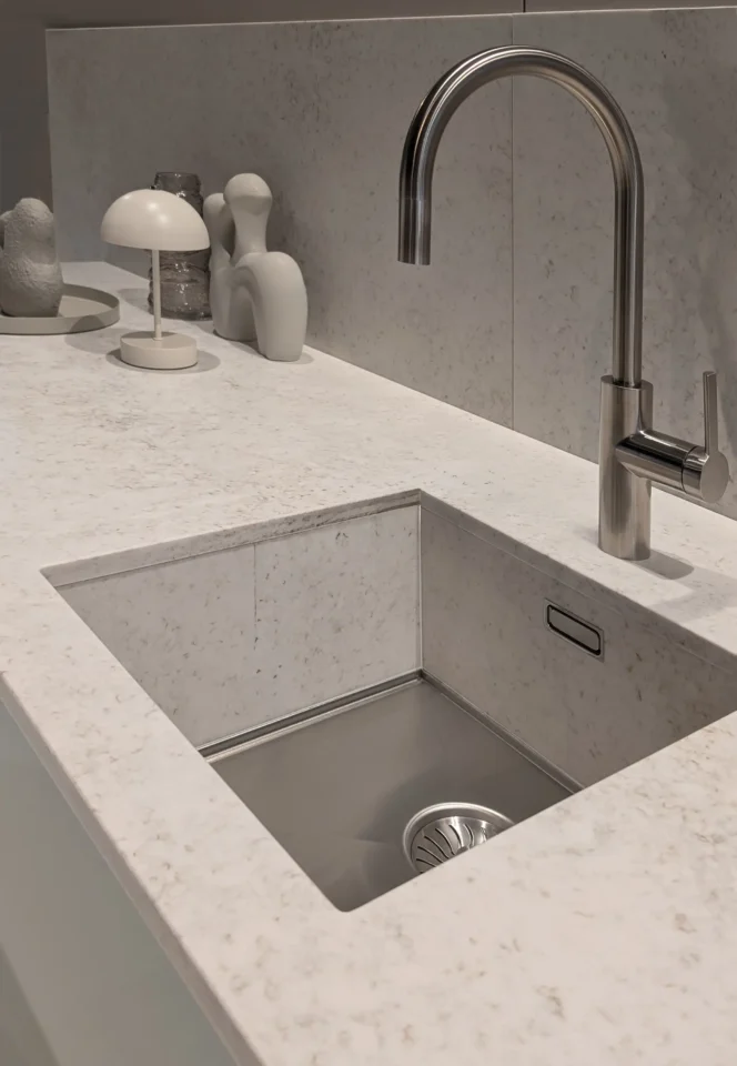 Ein moderner Waschbeckenbereich mit einer grauen Arbeitsplatte und einem rechteckigen Edelstahlspülbecken. Ein eleganter Wasserhahn, eine kleine weiße Lampe und dekorative Elemente befinden sich auf der Arbeitsplatte.