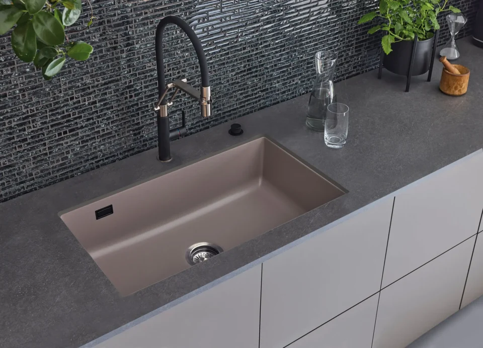 Eine moderne Küche mit grauer Arbeitsplatte und rechteckigem, beigem Spülbecken. Darüber befindet sich ein schwarzer Wasserhahn. Auf der Arbeitsplatte stehen ein Glas, eine Karaffe und eine Pflanze vor einer dunklen Fliesenrückwand.