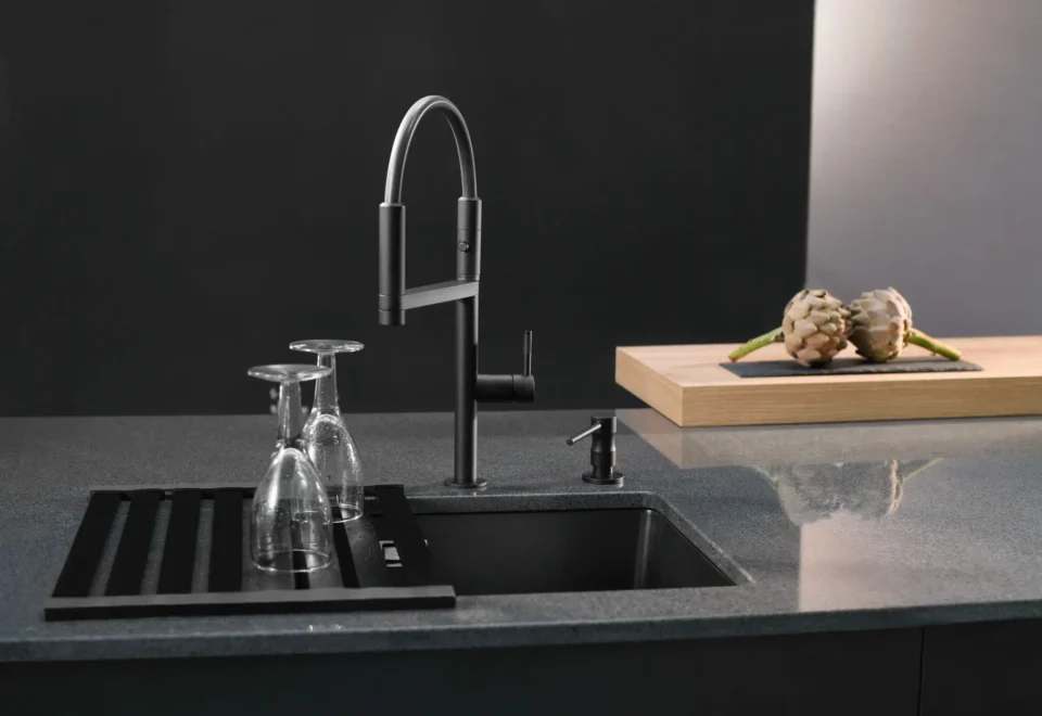 Eine moderne Küchenspüle in einer dunklen Arbeitsplatte mit einem hohen, gebogenen Wasserhahn. Auf einem Abtropfgestell stehen umgedrehte Gläser. Im Hintergrund liegt ein Schneidebrett mit zwei Artischocken.