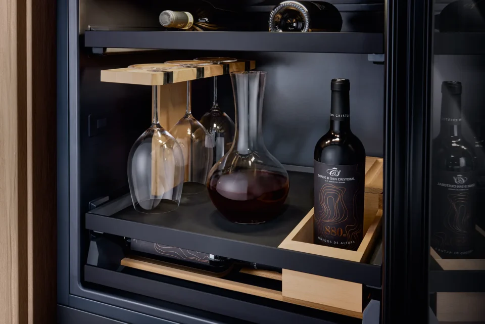 In einem Regal befinden sich mehrere Weinflaschen, ein Dekanter mit Rotwein und drei hängende Weingläser. Die Regalfächer sind dunkel und modern gestaltet.