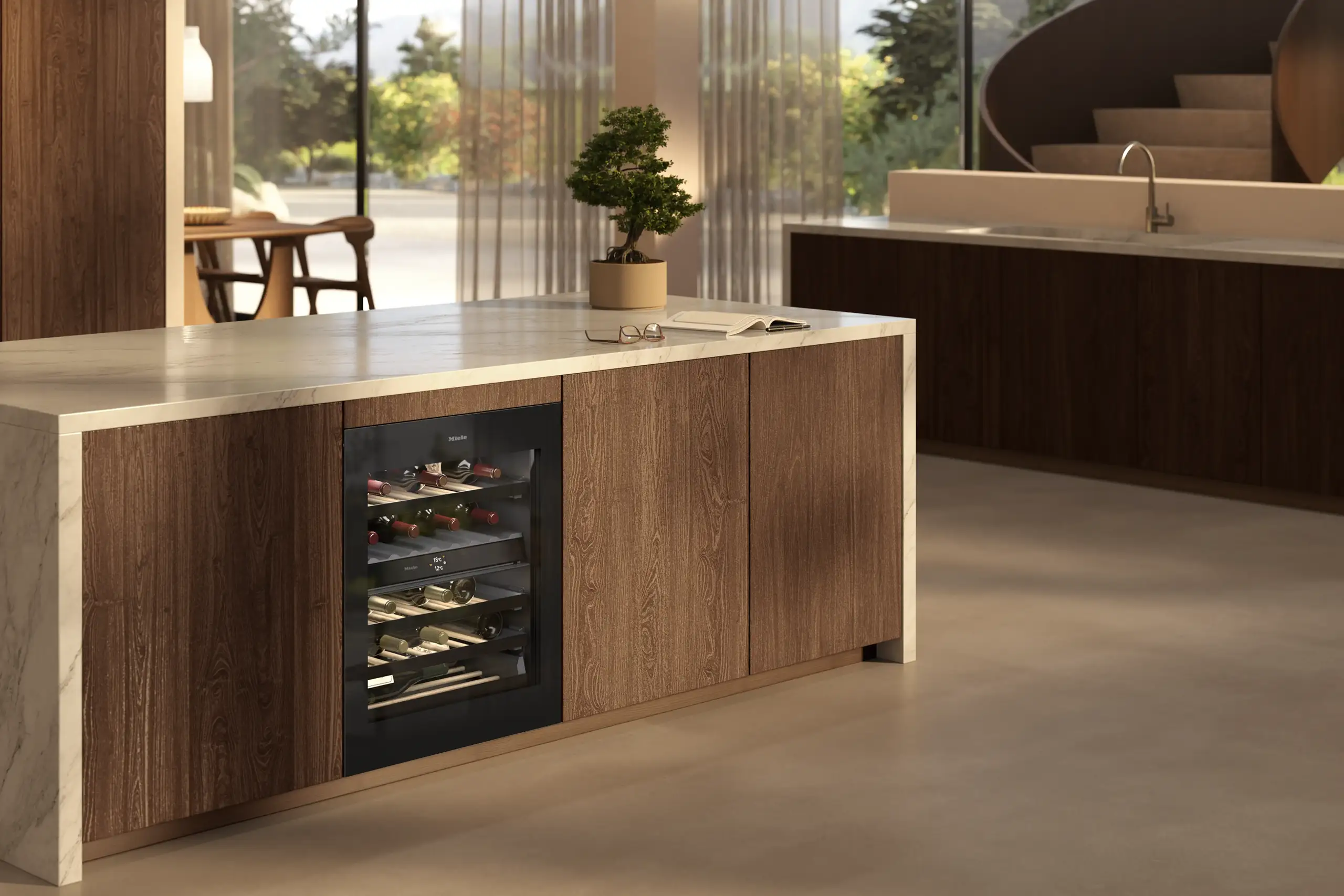Miele 20000212266 Moderne Küche mit Holzschränken und einer zentralen Insel mit eingebautem Weinkühler. Licht fällt durch große Fenster. Im Hintergrund befindet sich ein Tisch und eine Wendeltreppe. Ein Pflanzenarrangement schmückt die Insel.