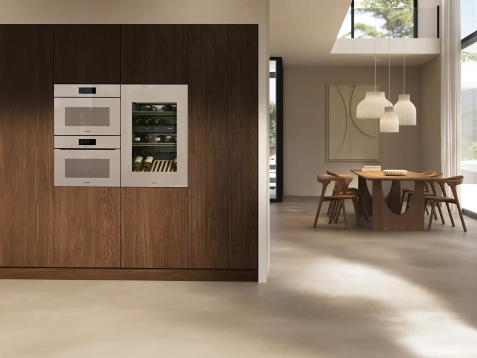 Moderne Küche mit eingebauten Elektrogeräten in einer Holzverkleidung. Rechts im Bild ein Essbereich mit einem Tisch und Stühlen. Hängende Lampen beleuchten den Raum durch große Fenster.