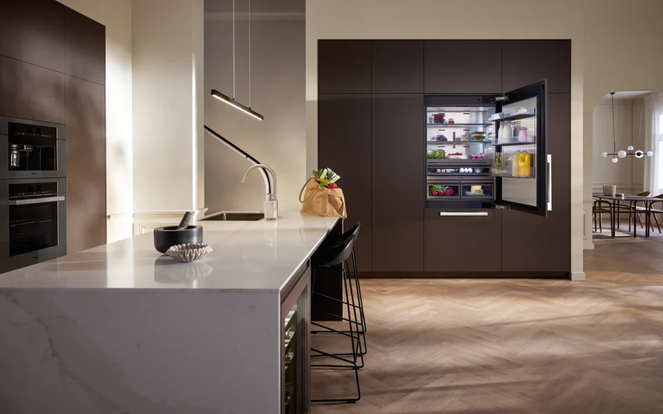 Moderne Küche mit Insel, ausgestattet mit dunklen Schränken und eingebautem Kühlschrank. Der Kühlschrank ist offen und zeigt Lebensmittel. Auf der Insel stehen Hocker, eine Schüssel und ein Brot auf einem Schneidebrett.