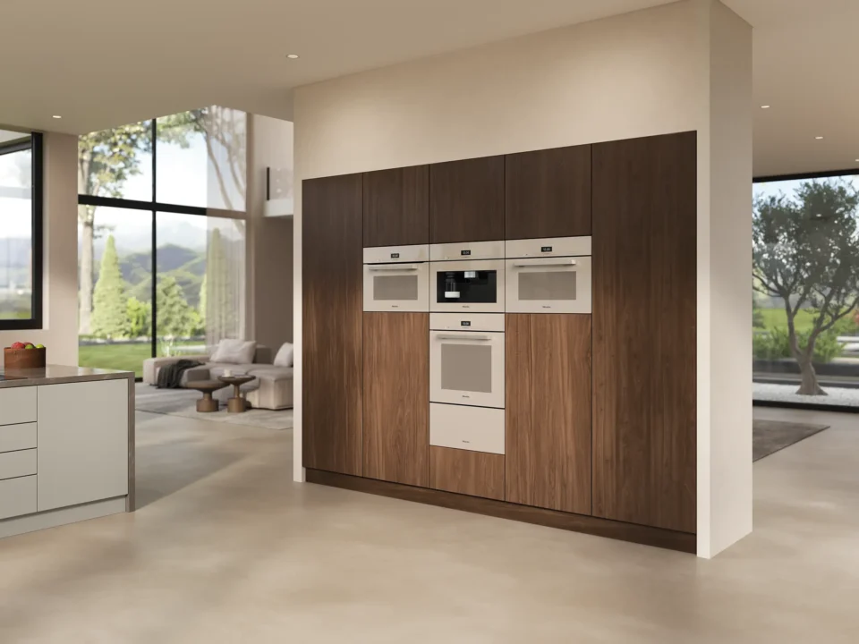 Moderne Küche mit Geräten in einer Holzverkleidung. Links sieht man weiße Arbeitsflächen und Schränke, im Hintergrund ein gemütlicher Wohnbereich mit Sofas und großen Fenstern, die den Blick auf Garten und Bäume freigeben.