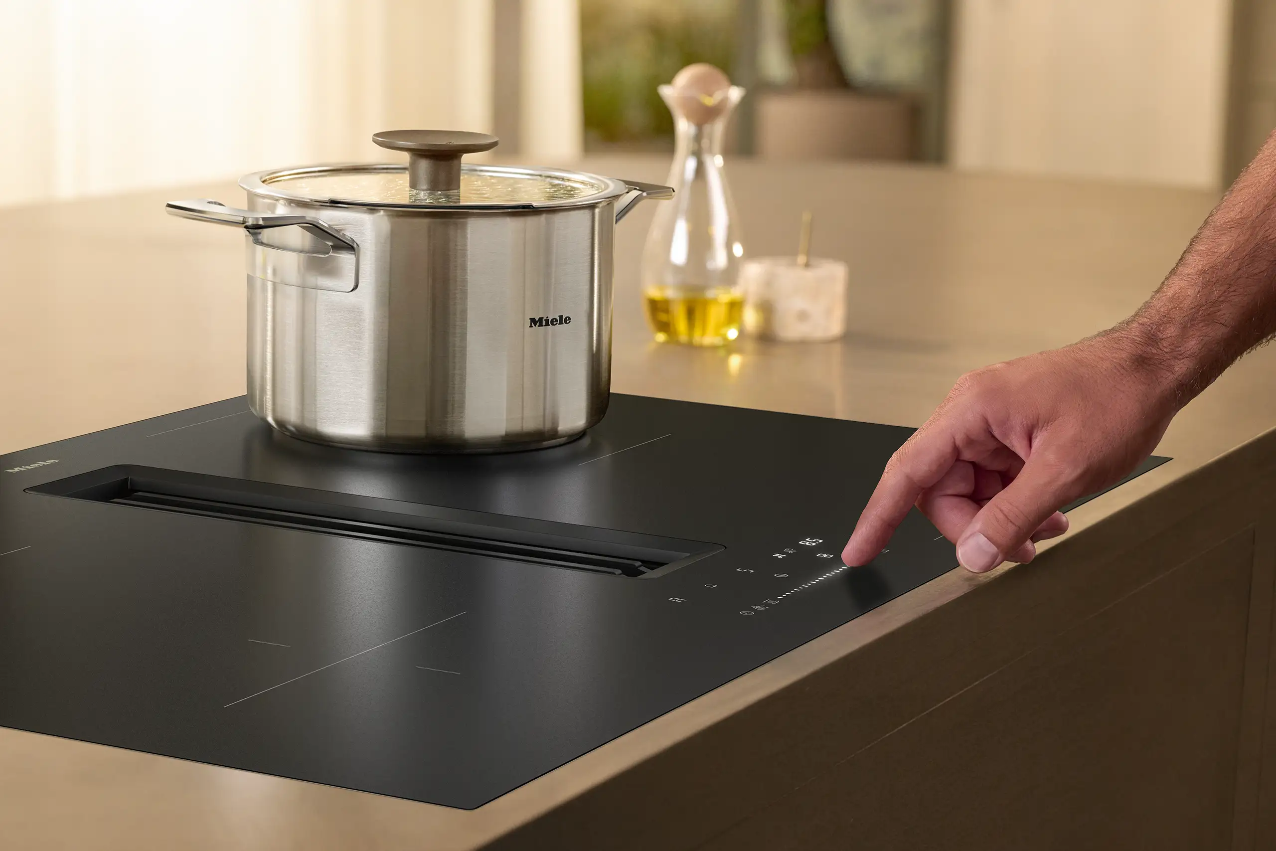 Miele 20000455690 Ein Finger bedient ein modernes, schwarzes Kochfeld mit einem Touch-Display. Auf dem Kochfeld steht ein großer Topf mit Deckel. Im Hintergrund sind eine Flasche Öl und ein Gewürzglas auf einer Arbeitsplatte zu sehen.