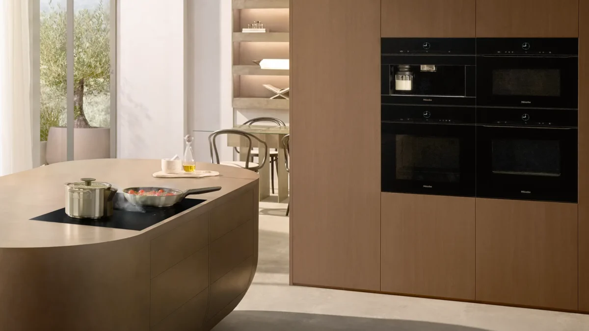 Miele 20000455698 Moderne Küche mit heller Holzverkleidung. Links ein abgerundeter Küchenblock mit Herdplatten und Töpfen. Rechts eingebauter Schrank mit vier schwarzen Geräten. Hinten Fenster und Regal mit Dekor. Neutrale, helle Farbgestaltung.
