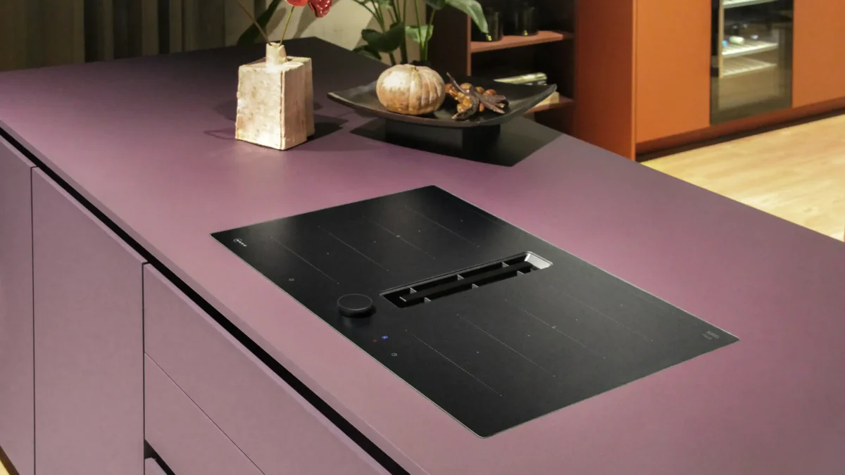 NEFF Matt Black Design V68PYX4B0 H4K Kitchen Lila Kücheninsel mit integriertem Kochfeld und schwarzem Dunstabzug. Auf der Arbeitsplatte stehen ein Pflanzenarrangement in einer Vase und ein flacher Teller mit Dekoration. Im Hintergrund sind Regale und ein Schrank sichtbar.