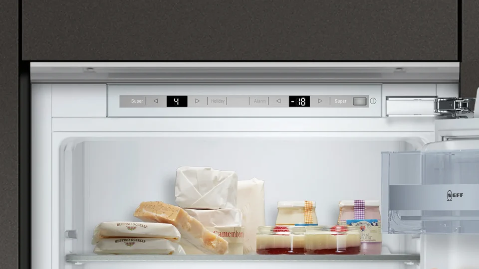 Ein offener Kühlschrank mit verschiedenen Lebensmitteln. Oben befinden sich digitale Bedienelemente mit Symbolen und Zahlen. Im Inneren sind Lebensmittel wie verpackter Käse, Brot und Gläser mit Joghurt zu sehen.