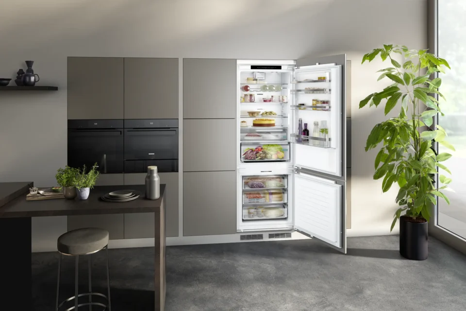 Ein moderner, geöffneter Kühlschrank in einer stilvollen Küche zeigt verschiedene Lebensmittel im Inneren. Links steht ein Tisch mit Gewürzen und einer Wasserflasche, rechts eine große grüne Pflanze. Dezente Beleuchtung ergänzt das Ambiente.