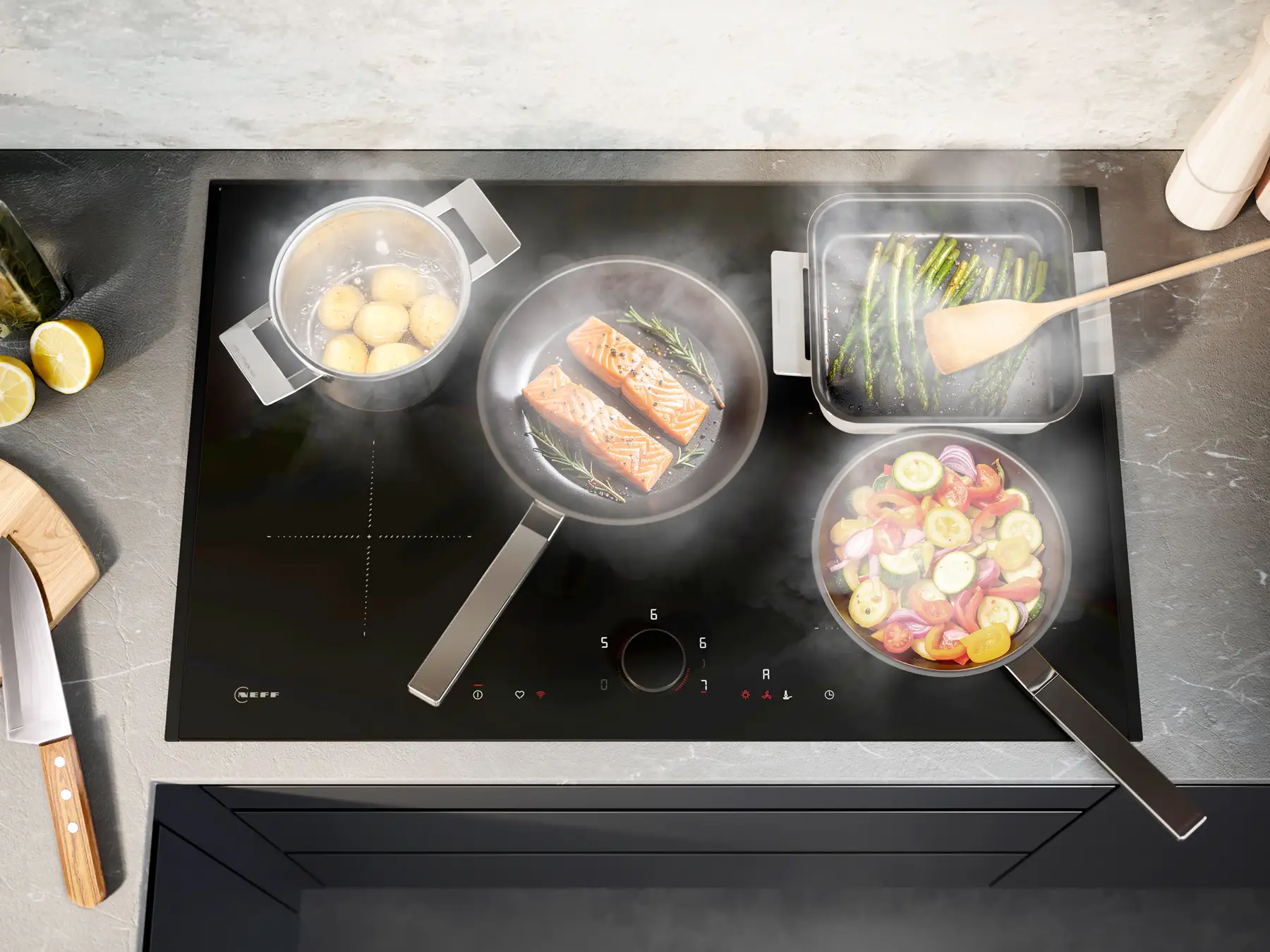 REU SV NEFF Matt Black Design Induction NEFF Collection Image 23 MattBlackDesign Ein schwarzes Induktionskochfeld mit vier Töpfen und Pfannen steht auf einer Arbeitsplatte. Darin sind Kartoffeln, Lachs mit grünem Gemüse, grüne Bohnen und buntes Gemüse mit einer Holzkelle zu sehen. Dampf steigt auf.