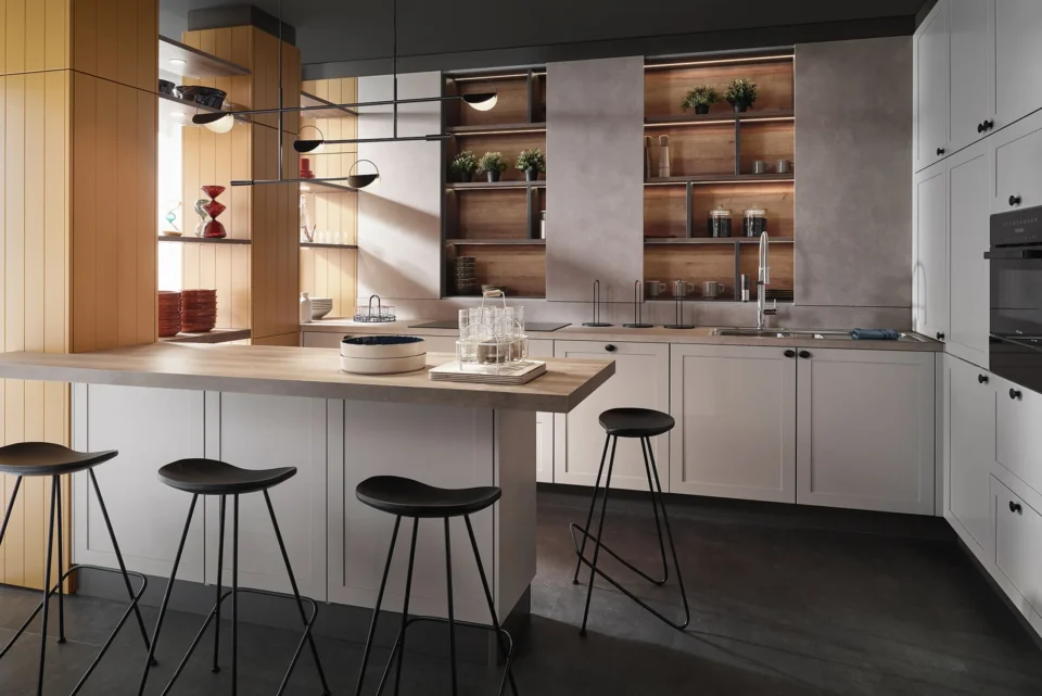 Moderne Küche mit grauen und weißen Schränken, einer zentralen Insel mit vier schwarzen Hockern, offenen Regalen mit Pflanzen und Geschirr, Spüle und eingebautem Ofen. Dezente Beleuchtung und Holzakzente verleihen Gemütlichkeit.