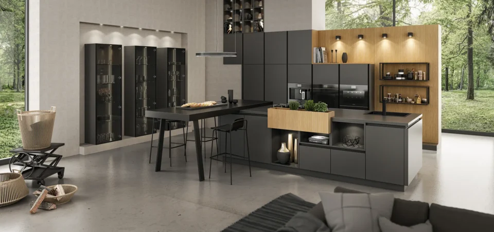 Moderne Küche mit grauen Schränken, integriertem Ofen und Kaffeemaschine. Ein zentraler Tisch mit Barhockern steht im Raum. Offene Regale mit Beleuchtung und Dekoration. Große Fenster mit Blick auf einen grünen Wald.