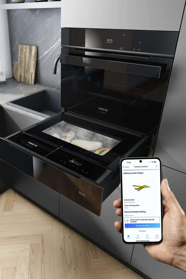 Ein moderner, schwarzer Ofen in einer Kücheneinrichtung. Eine Hand hält ein Smartphone mit einer Kochassistent-Anwendung. Der Ofen hat zwei Auszüge, eine Glasscheibe zeigt das Innere mit einem Gericht.