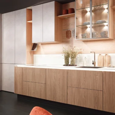 Moderne Küche mit Holzfronten, weißer Arbeitsfläche und eingebauten Elektrogeräten. Oberschränke und offene Regale mit Tassen und Vasen. Beleuchtung betont die Glasvitrine. Dunkler Boden und helle Wände vervollständigen das Design.