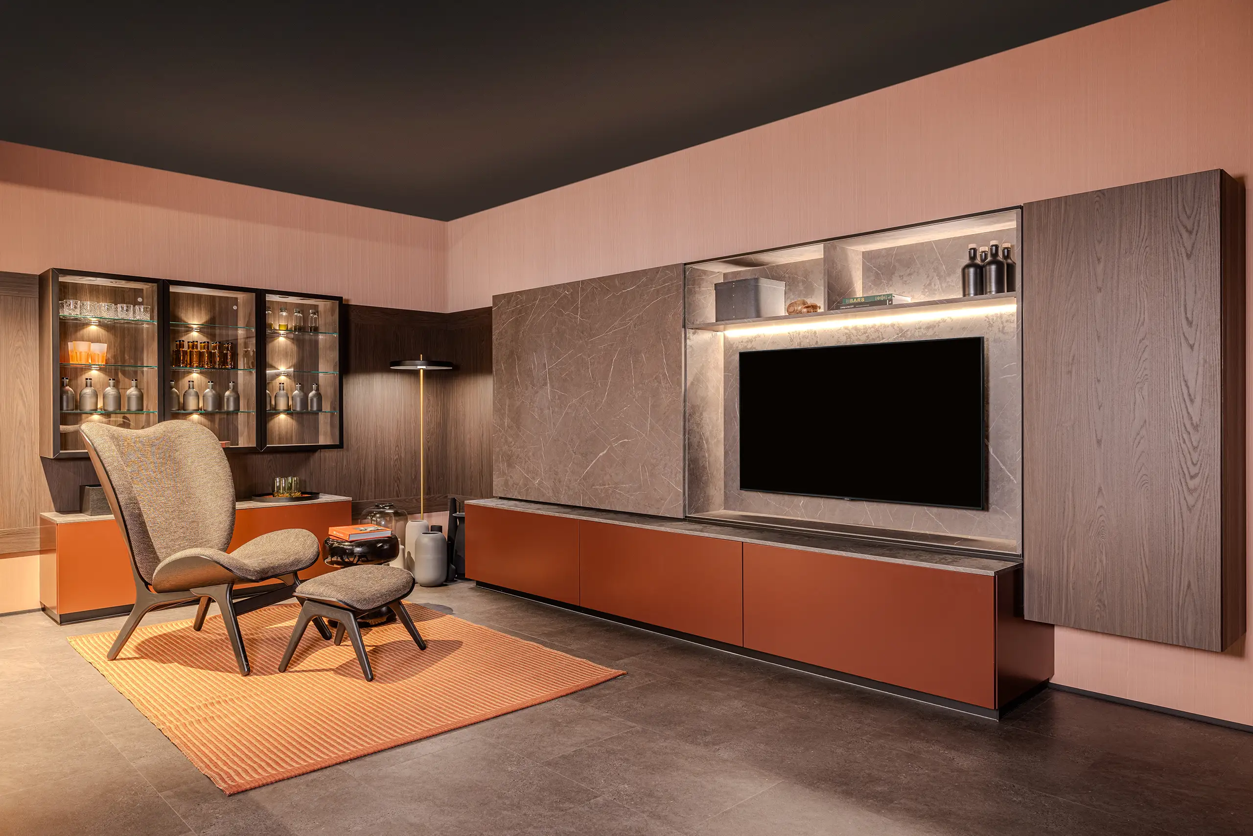 Ein modernes Wohnzimmer mit grauem Boden, einem großen Fernseher in einem Regal, umgeben von braunen und grauen Holzmöbeln. Links steht ein Stuhl mit Ottoman auf einem orangefarbenen Teppich. Hinterleuchtet sind Gläser und Flaschen in einem Schrank.