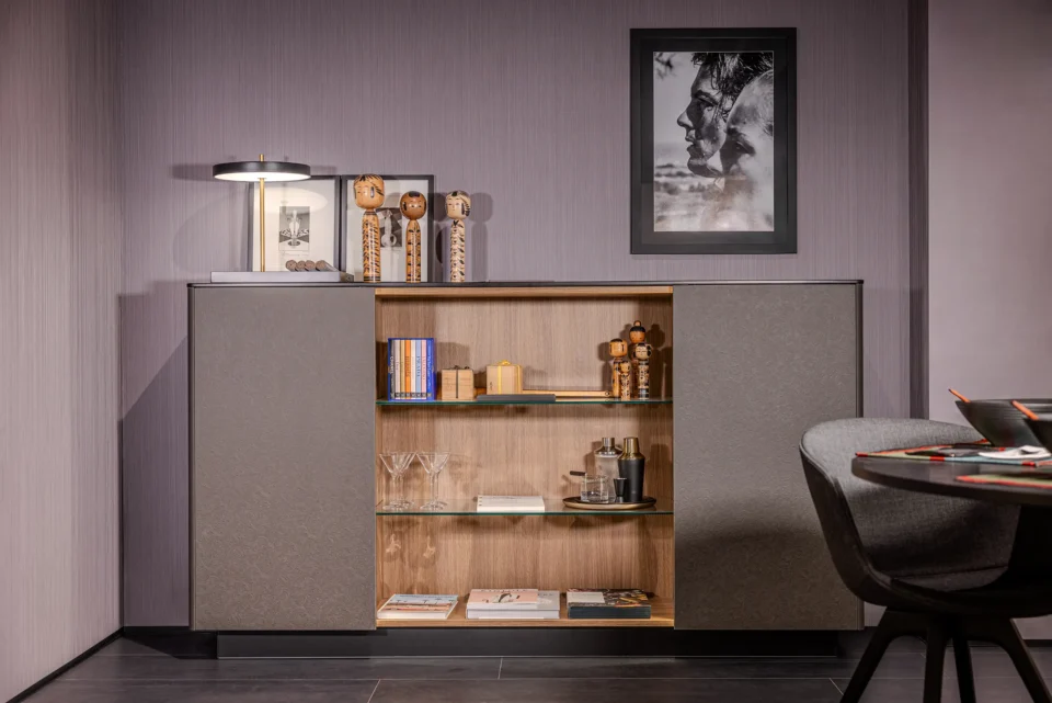 Ein Wohnzimmer mit einem modernen Sideboard an einer grauen Wand. Darauf stehen eine Lampe und Dekorationen. Im offenen Fach sind Bücher und Gläser. Oben hängt ein gerahmtes Bild. Rechts ist ein Teil eines Stuhls erkennbar.