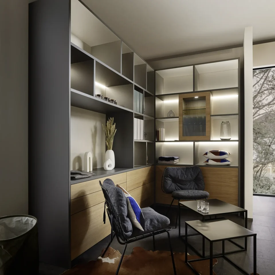 Modernes Wohnzimmer mit Wandregalen, Schränken und Deko-Objekten. Zwei schwarze Stühle und ein Beistelltisch stehen auf einem dunklen Teppich. Ein großes Fenster bietet Blick auf Bäume draußen. Dezente Beleuchtung setzt Akzente.