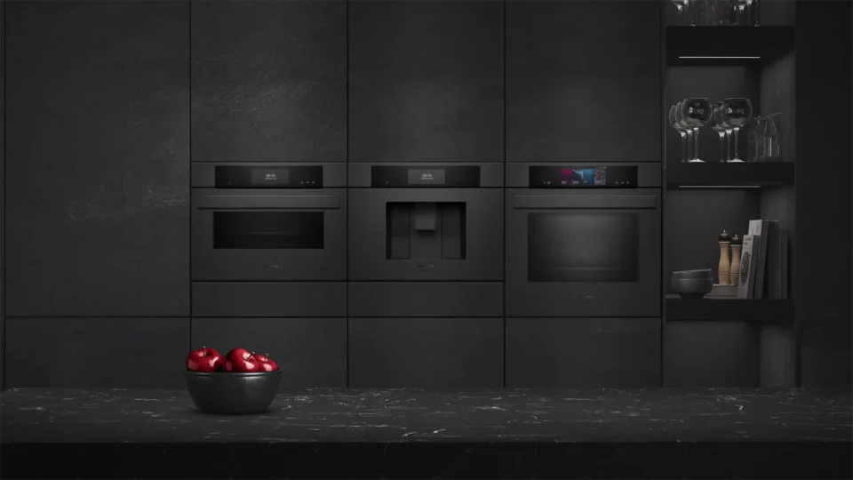 Eine moderne Küche mit schwarzen Einbaugeräten: ein Dampfgarer, eine Kaffeemaschine und ein Backofen. Im Vordergrund steht eine schwarze Schale mit roten Äpfeln auf einer schwarzen Arbeitsplatte. Rechts sind Gläser in einem Regal zu sehen.