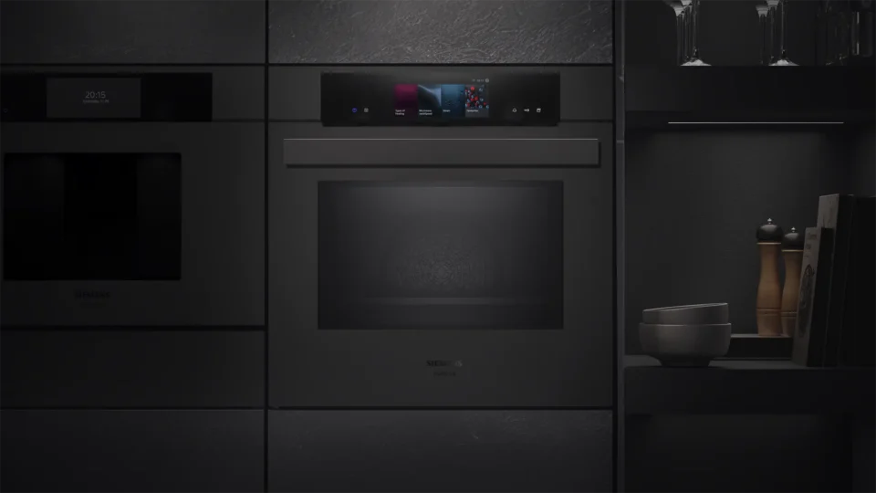 Ein moderner, schwarzer Einbau-Backofen mit digitalem Display und Touchscreen in einer schwarzen Küchenzeile ist zu sehen. Rechts steht eine Schale auf einem Regal, daneben sind einige Gläser.