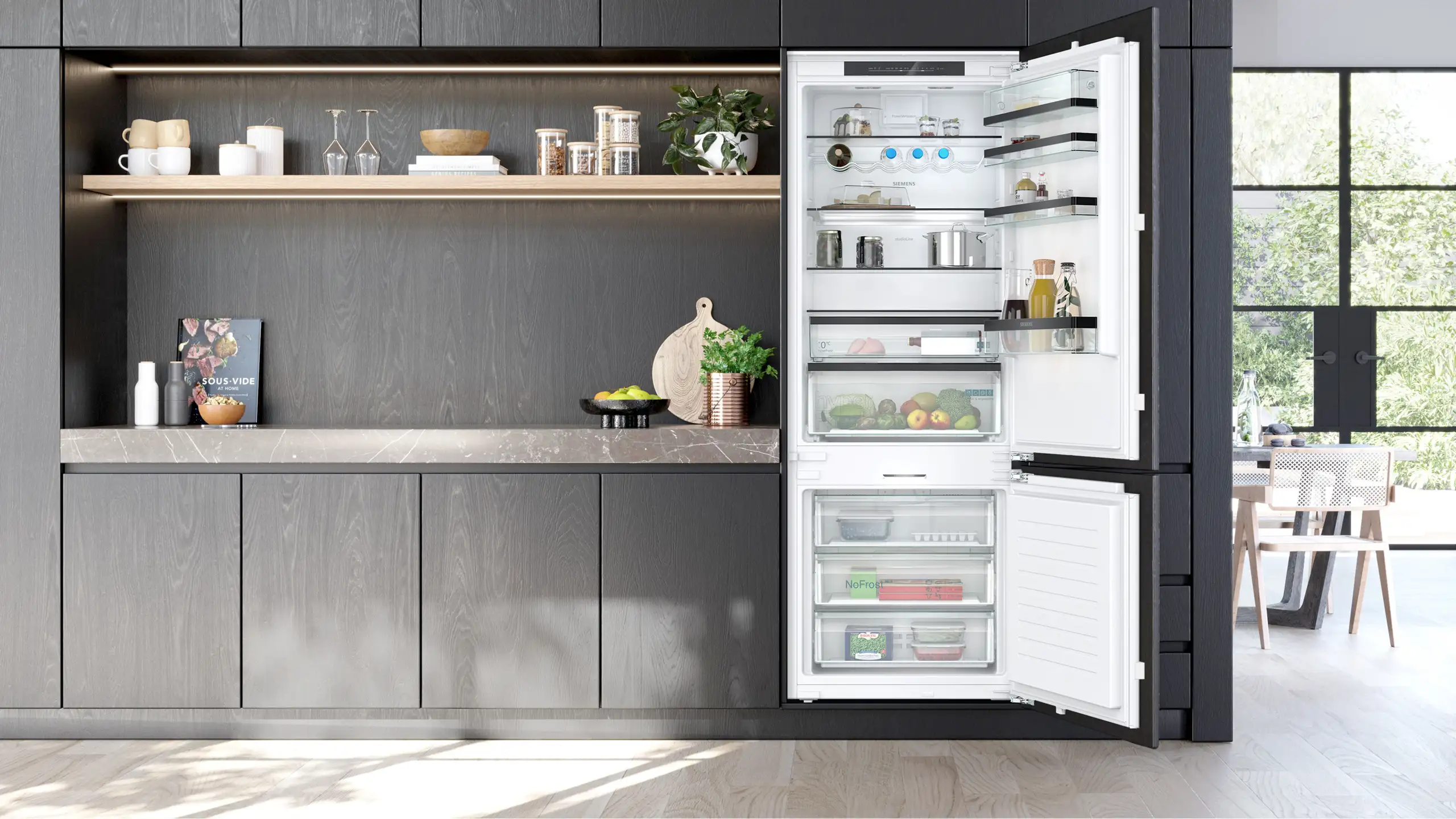 Eine moderne Küche mit dunklen Holzschränken und offenem Regal. Rechts ist ein geöffneter Kühlschrank mit Lebensmitteln und Getränken gefüllt. Tageslicht strömt von einem großen Fenster auf der rechten Seite herein.