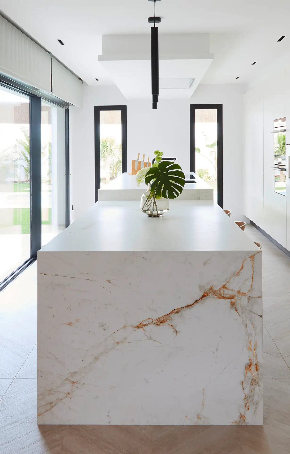 Moderne Küche mit weißer Marmorinsel, darauf eine Vase mit grünen Blättern. Große Fenster lassen viel Licht herein. Klare Linien und eine minimalistische Gestaltung dominieren den Raum.