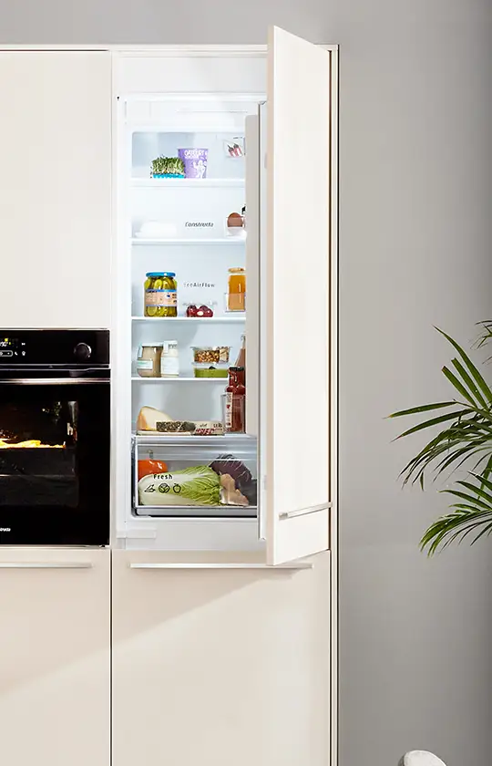 De Koeoek Marken List Constructa Ein geöffneter Einbaukühlschrank zeigt mehrere Regale mit Lebensmitteln wie Früchten, Saft und Soßen. Links daneben ist ein geschlossener Backofen. Im Vordergrund steht eine Pflanze mit grünen Blättern.