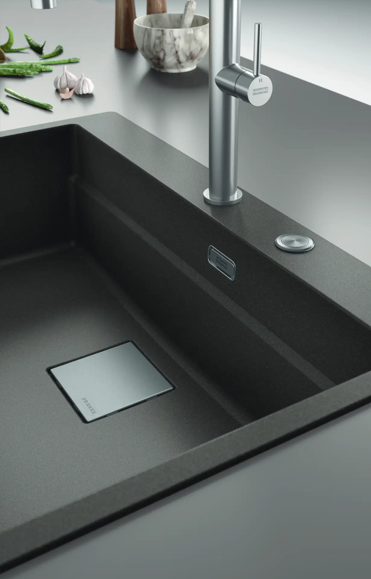 De Koeoek Marken List Franke Ein moderner, schwarzer Küchenspülbeckenbereich mit einem rechteckigen Abfluss. Daneben sind ein silberner Wasserhahn, eine Ablauffernbedienung und auf der Arbeitsfläche grüne Bohnen und Knoblauchzehen zu sehen.