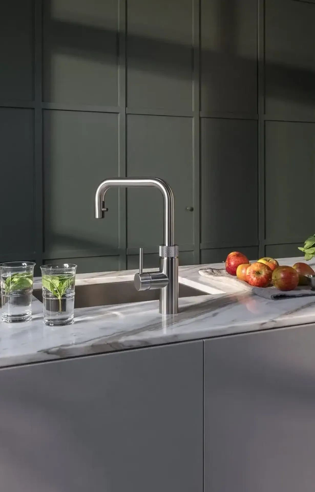 De Koeoek Marken List Quooker Eine Person bedient einen modernen schwarzen Wasserhahn an einem Edelstahlspülbecken. Aus dem Wasserhahn fließt Wasser. Ein roter Ring am Griff deutet auf eine Temperaturregelung hin.
