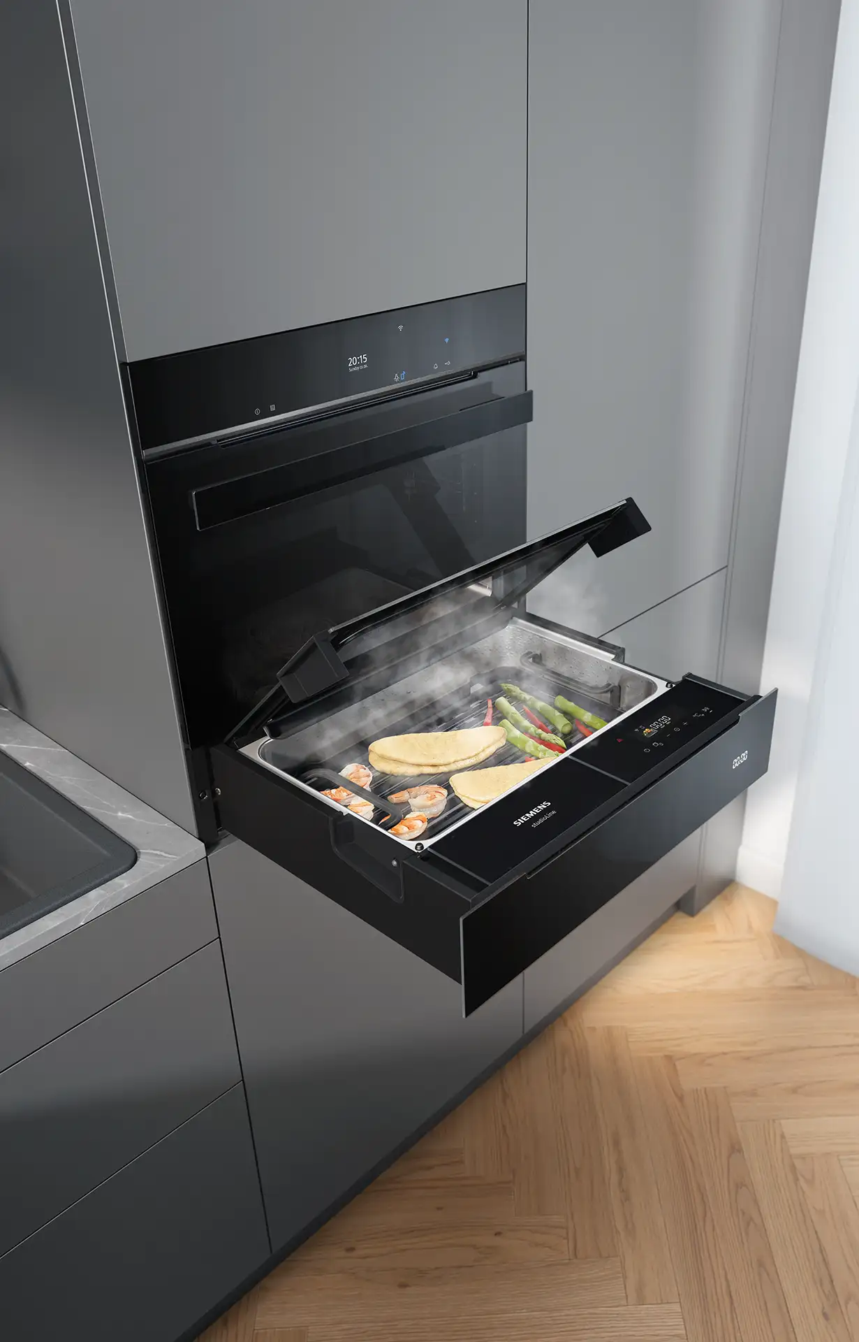 De Koeoek Marken List Siemens Ein Dampfgarer öffnet sich seitlich in einer modernen Küchenkombination. Innen liegen verschiedene Gemüsesorten und zwei Tortillas auf einem Tablett. Dampf steigt auf, die Küche hat graue Schränke und einen Holzboden.