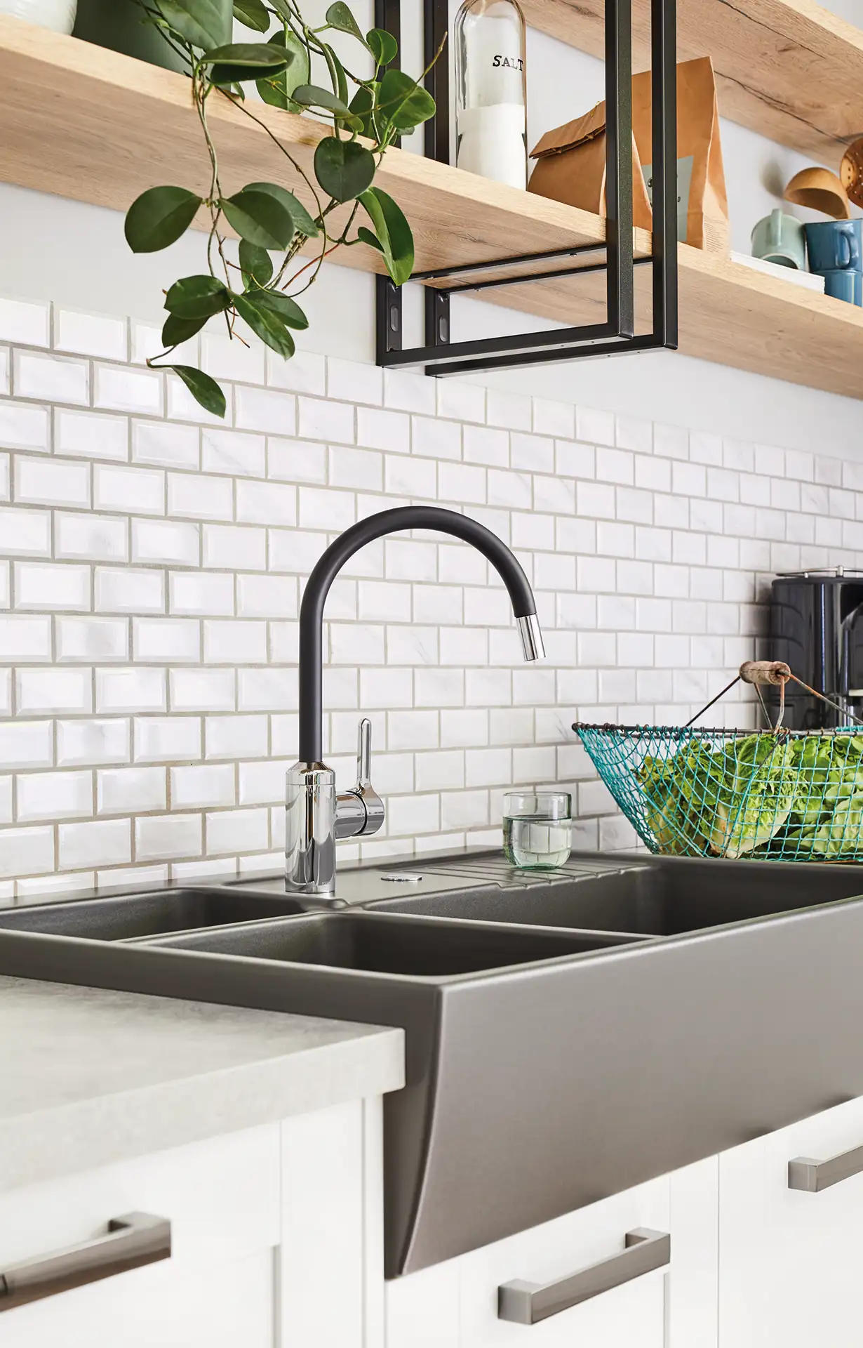 De Koeoek Marken List Systemceran Küche mit weiß gefliester Rückwand, grauem Spülbecken und schwarzem Wasserhahn. Darüber schwebende Regale mit Pflanzen, Tassen und Schalen. Rechts ein Drahtkorb mit grünem Gemüse.
