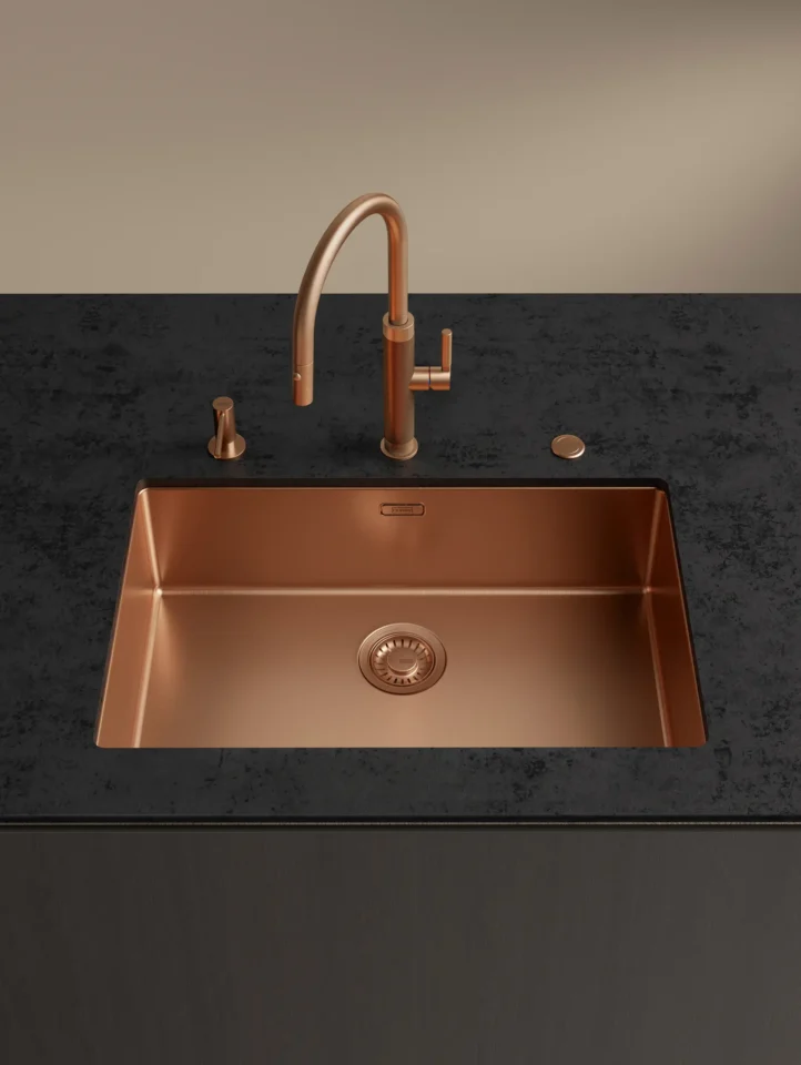 Küchenarbeitsplatte aus dunklem Stein mit eingebautem, rechteckigem Kupferspülbecken und passendem Wasserhahn. Links ein Seifenspender, rechts ein Abflussdeckel. Hintergrund in sanften Beige- und Sandtönen.