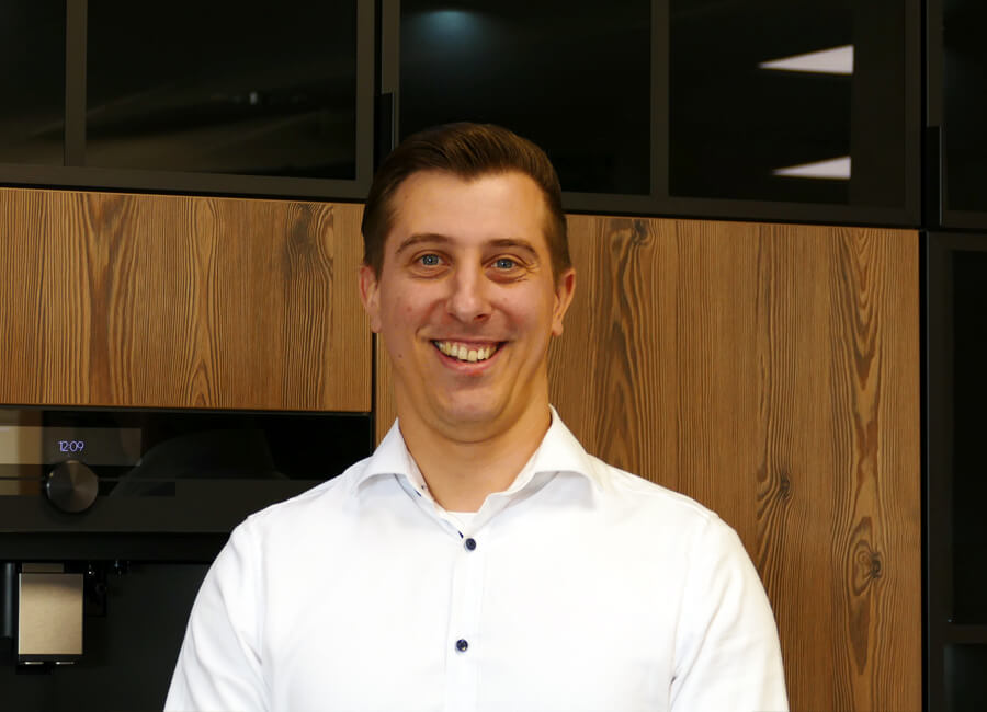 Eine Person mit kurz geschnittenem Haar trägt ein weißes Hemd und steht lächelnd in einer Küche vor einem Holzschrank mit schwarzen Akzenten. Daneben sind Teile eines schwarzen Küchengeräts sichtbar.
