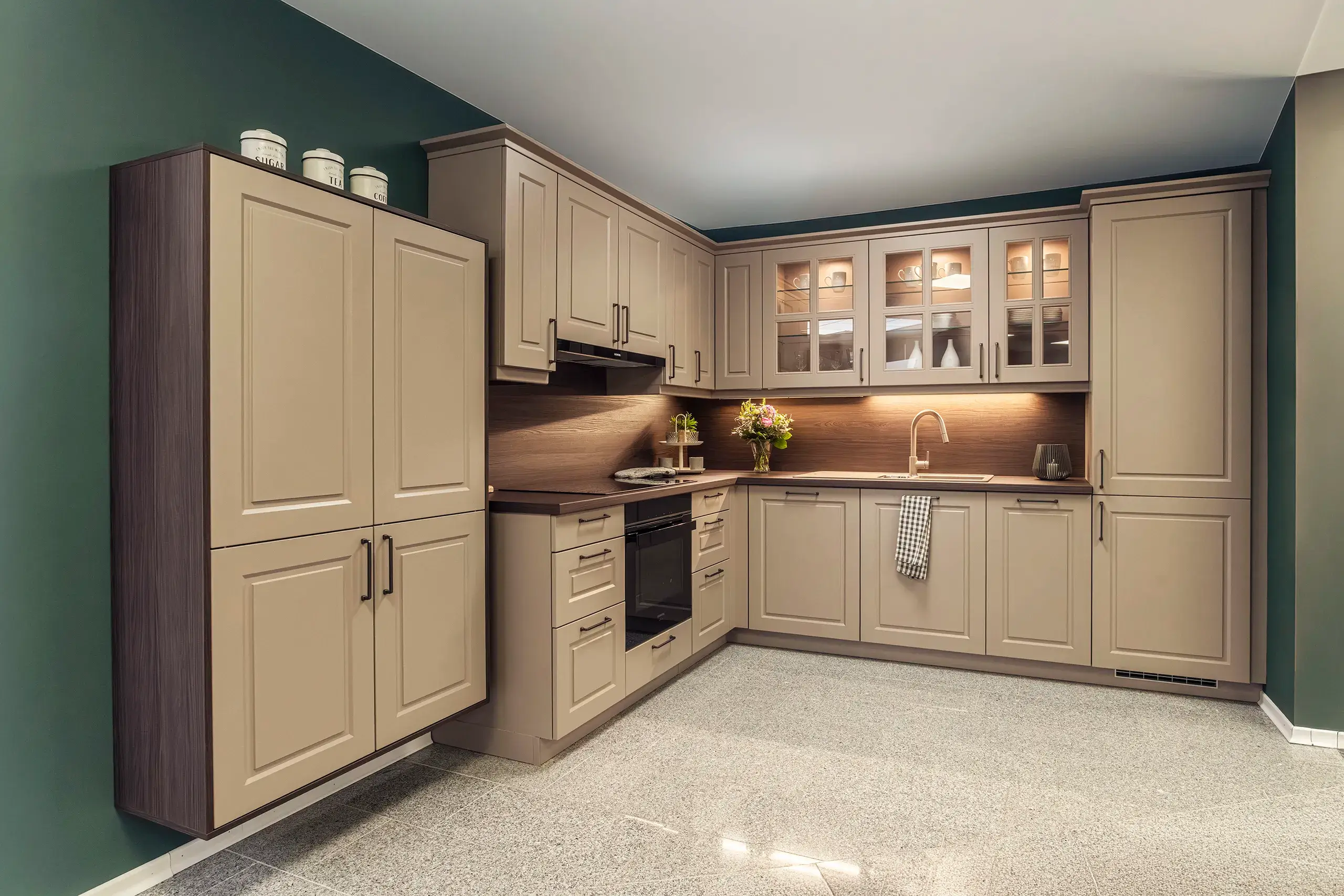 Kuechenstudio De Koeoek Datenschutz Moderne Küche mit L-förmigen beigefarbenen Schränken, schwarzem Herd und Spüle. Oberhalb sind beleuchtete Glasvitrinen. Links stehen Vorratsbehälter auf einem hohen Schrank. Fußboden in neutralem Ton.