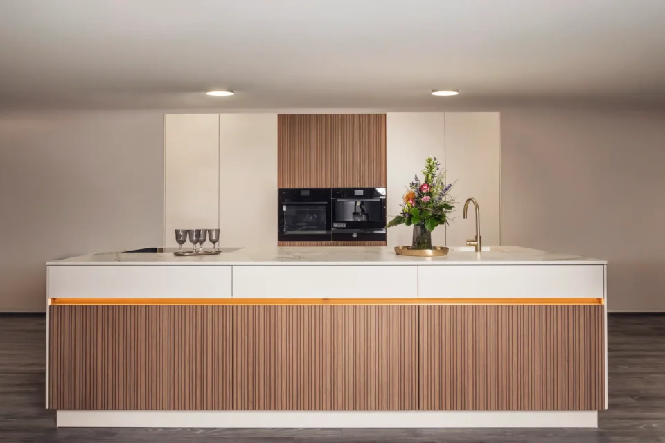Moderne Küche mit Insel in der Mitte. Die Kücheninsel hat eine beige und hölzerne Verkleidung mit einer goldenen Wasserarmatur und einem Blumenstrauß. Im Hintergrund sind ein eingebauter Backofen und Schränke zu sehen.