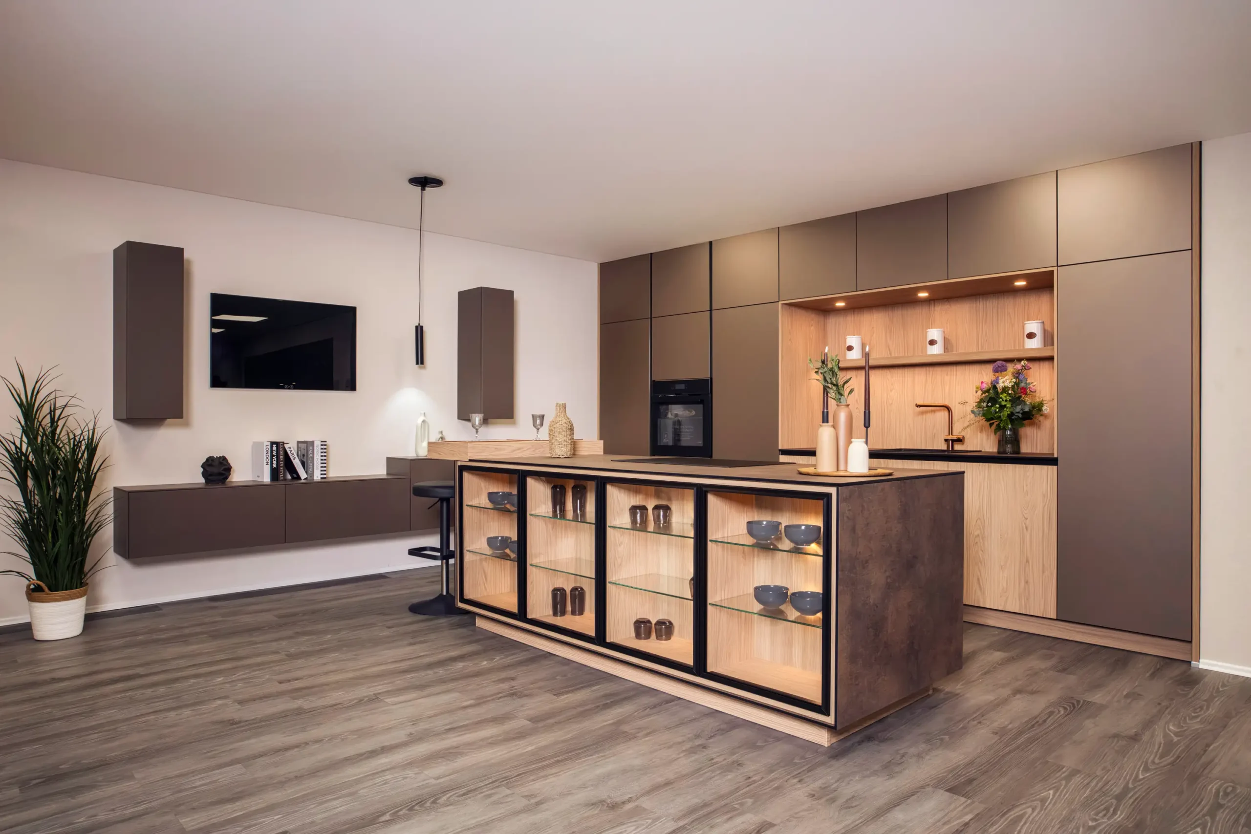 Kuechenstudio De Koeoek Designkuechen 2 Moderne Küche mit Kochinsel, dunkelbraunen Schränken und Holzakzenten. Hängeschränke und Regal mit Dekorationen. Linke Wand mit schwebendem Schrank und Fernseher, rechte Seite mit eingebautem Ofen. Holzlaminatboden.