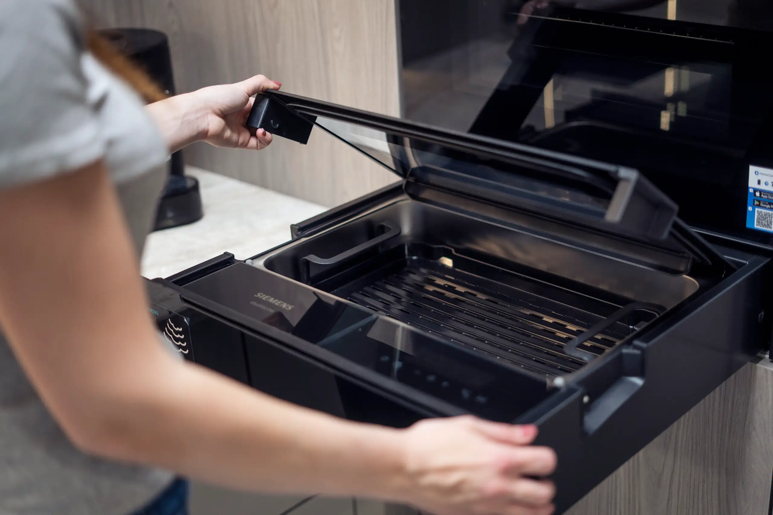 Kuechenstudio De Koeoek Einbaugeraete 4 Eine Person öffnet die Schublade eines Dampfgarers in einer modernen Küche. Die Schublade enthält einen schwarzen Grillrost. Die Person trägt ein graues Oberteil, und im Hintergrund sind Küchenschränke zu sehen.
