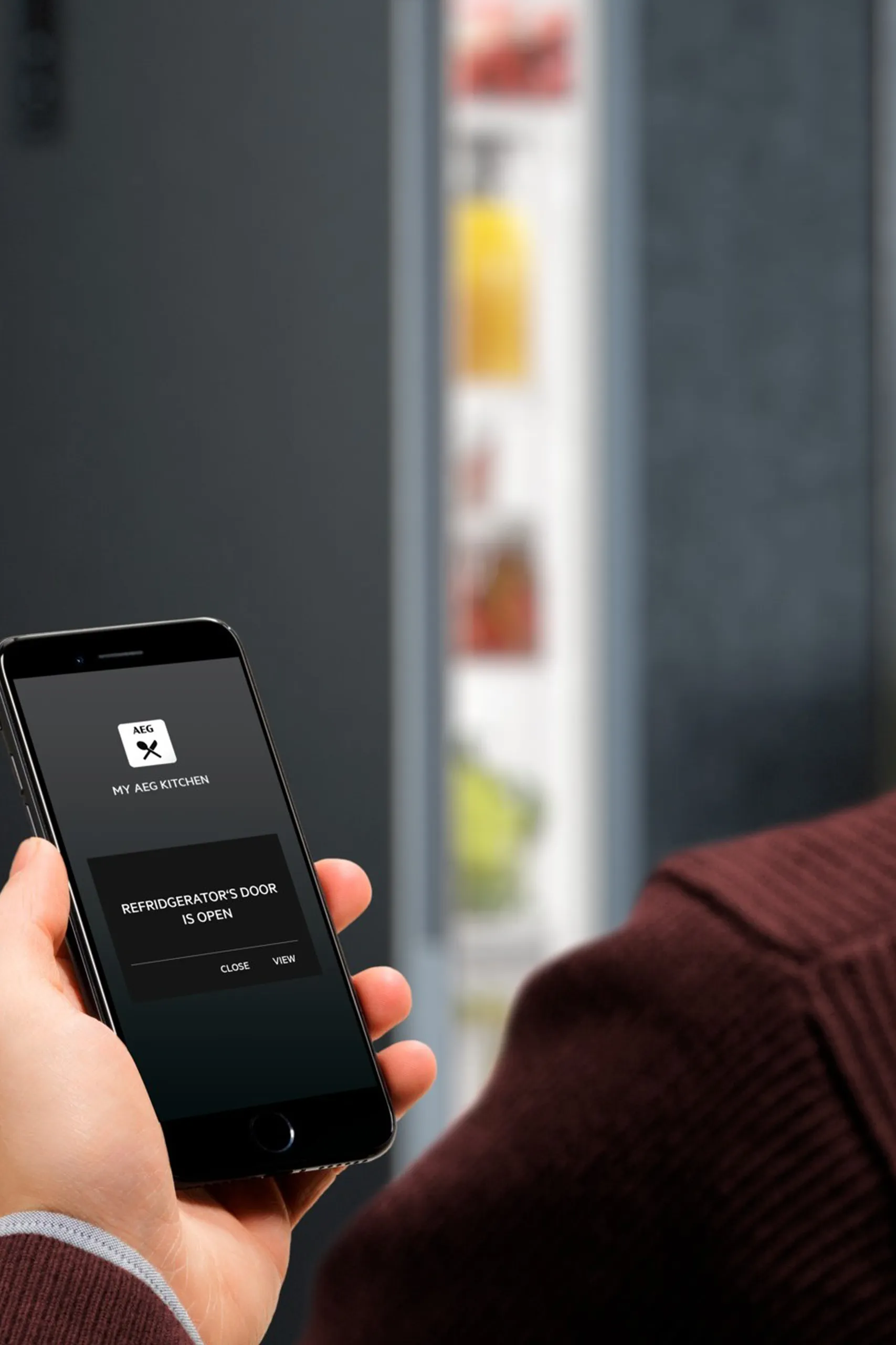 Eine Person hält ein Smartphone, das eine Nachricht anzeigt: „Kühlschranktür ist offen“. Im Hintergrund ist ein leicht geöffneter Kühlschrank mit unscharfen Objekten zu sehen.