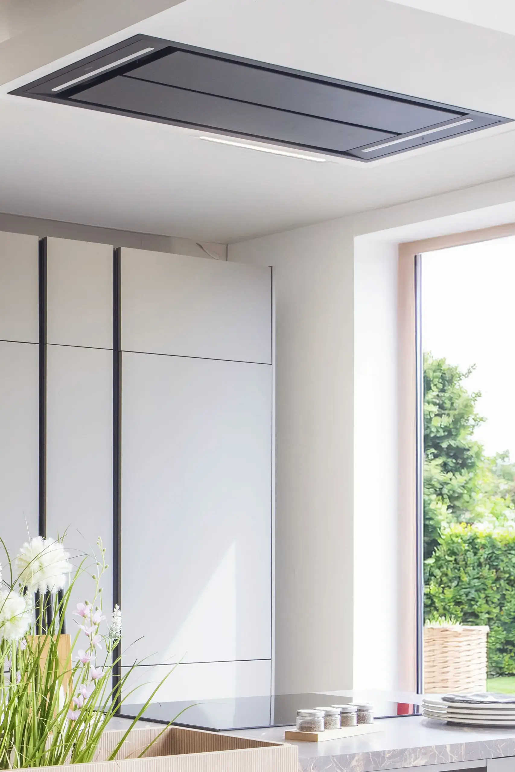 Moderne Küche mit weiß-grauen Schränken und einem großen Fenster, das einen Blick ins Grüne bietet. An der Decke ist eine flache, rechteckige schwarze Dunstabzugshaube angebracht. Im Vordergrund stehen Blumen.
