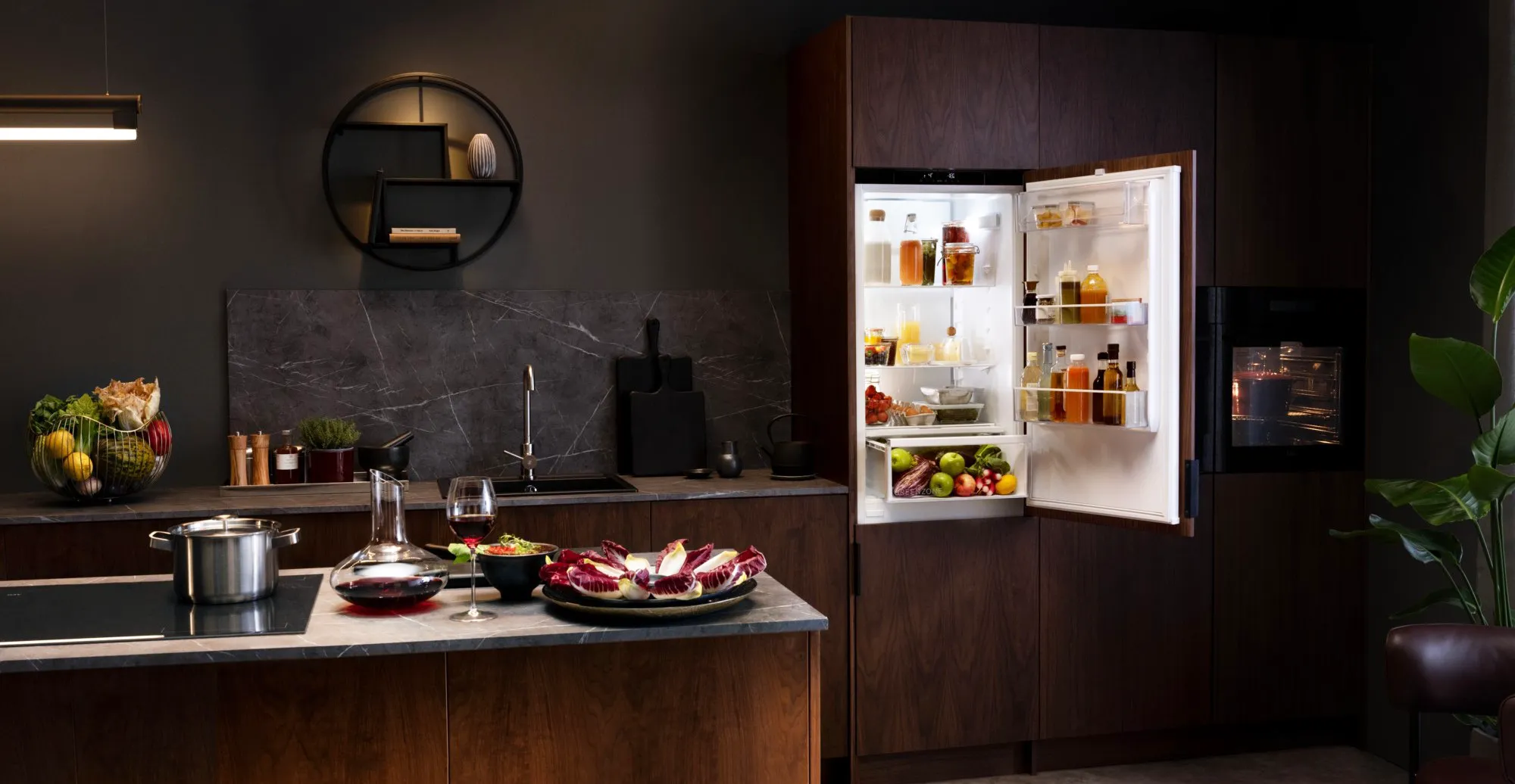 Moderne Küche mit dunklen Holzschränken, einer Arbeitsplatte mit Gemüseschale und Weingläsern. Ein offener Kühlschrank zeigt Lebensmittel und Getränke. Wandregale mit Dekoration und eine Zimmerpflanze ergänzen den Raum.