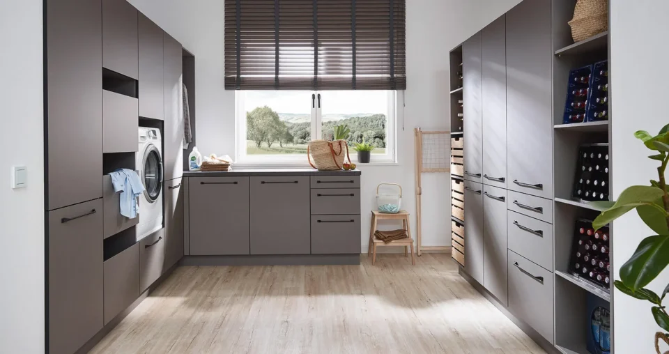 Moderner Hauswirtschaftsraum mit grauen Schränken und Regalen, einer Waschmaschine, einem großen Fenster mit Jalousien, hellem Holzboden, einem kleinen Stuhl und Pflanzen. Draußen sind Bäume zu sehen.