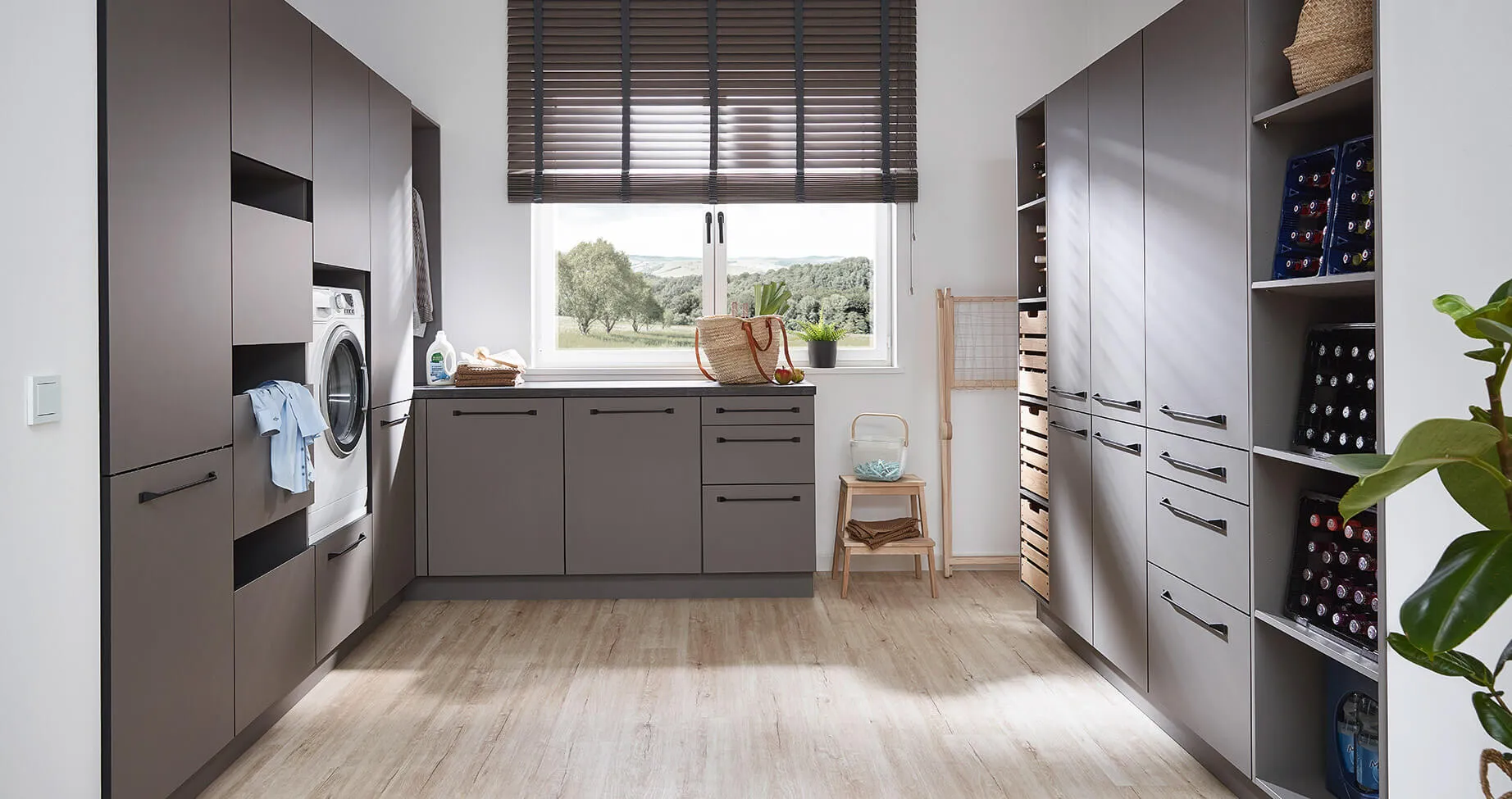 Kuechenstudio De Koeoek Hauswirtschaftsraum Ordnung 02 Moderner Hauswirtschaftsraum mit grauen Schränken und Regalen, einer Waschmaschine, einem großen Fenster mit Jalousien, hellem Holzboden, einem kleinen Stuhl und Pflanzen. Draußen sind Bäume zu sehen.