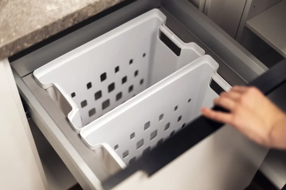 Ein offener Küchenschrank mit zwei herausnehmbaren weißen Mülltrennbehältern, die seitliche Griffe und quadratische Lüftungslöcher haben. Eine Hand bewegt die Schranktür.