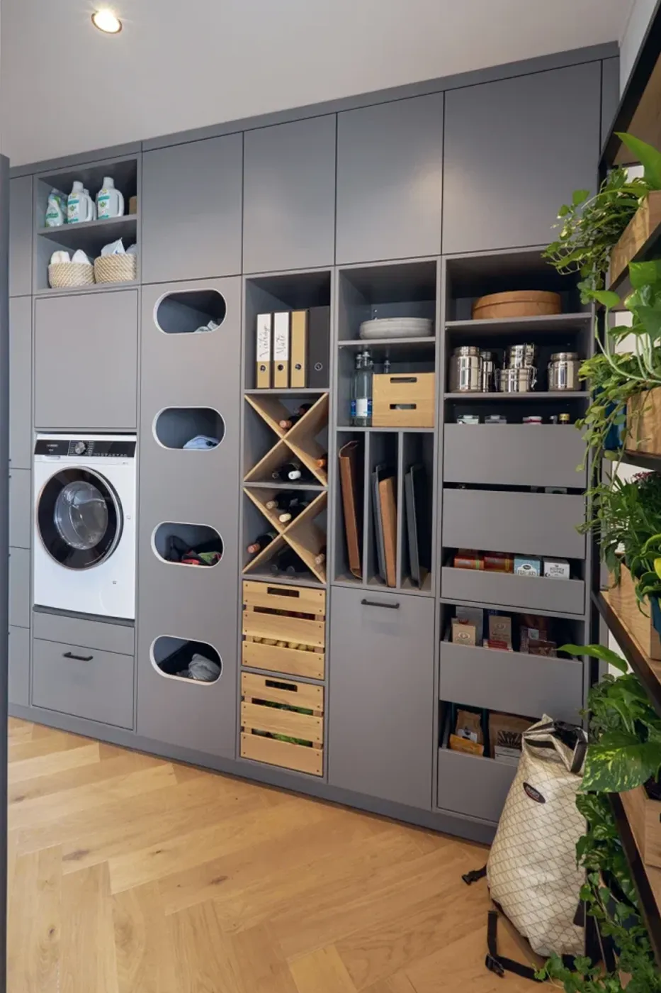 Kuechenstudio De Koeoek Hauswirtschaftsraum Bild eines modernen Hauswirtschaftsraums mit grauen Schränken. Links befindet sich eine Waschmaschine mit integriertem Trockner. Rechts sind Regale mit verschiedenen Fächern und Boxen für Aufbewahrung und Organisation sichtbar, daneben ein Korb und Pflanzen.