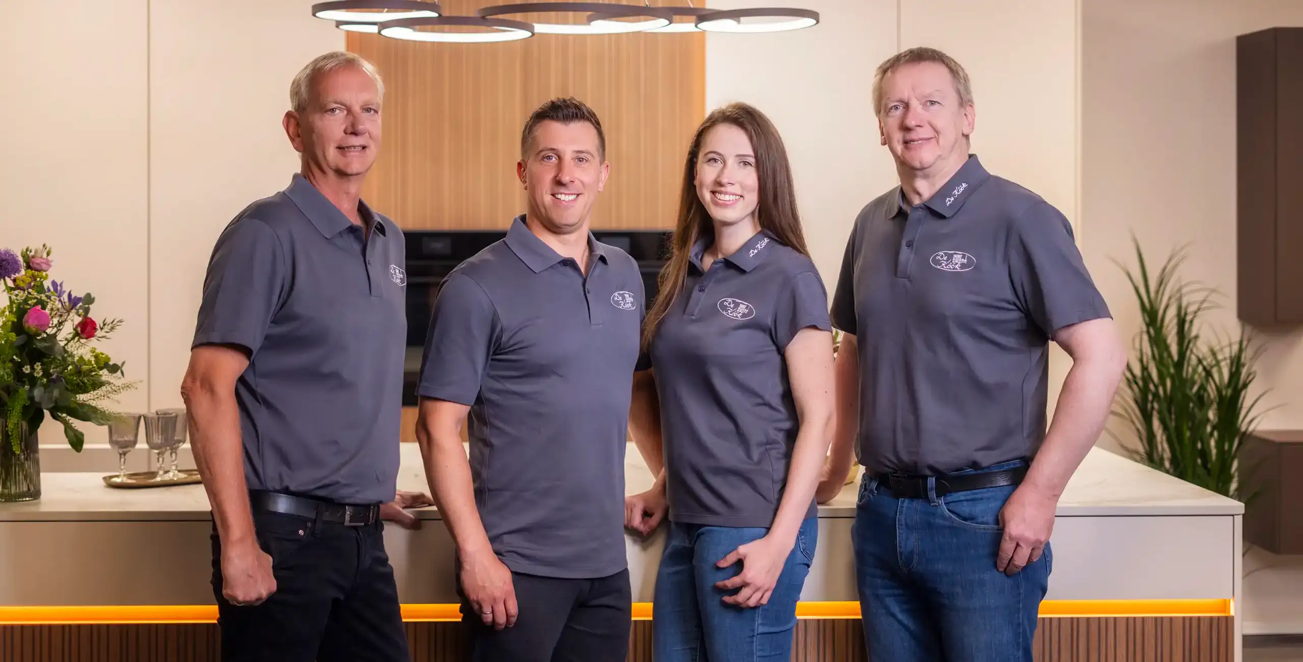 Vier Personen in grauen Poloshirts stehen in einer modernen Küche. Im Hintergrund sind Holzschränke und ein Blumenarrangement sichtbar. Alle lächeln in die Kamera und die Beleuchtung ist hell und einladend.