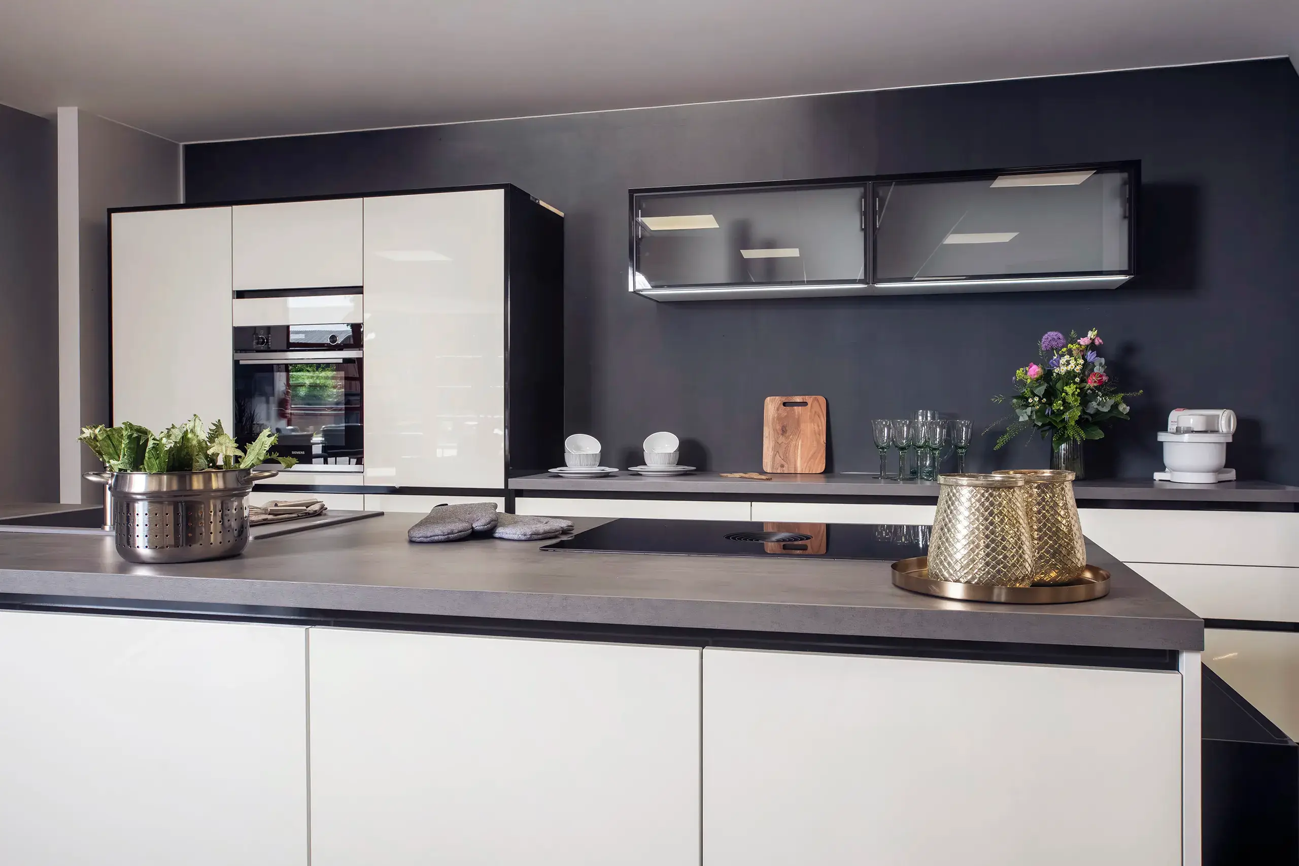 Kuechenstudio De Koeoek Impressum Moderne Küche mit weißen Hochglanz-Schränken, dunkler Wand, integriertem Backofen und Herd auf der Kücheninsel. Links ein Sieb mit Kräutern, rechts ein Tablett mit goldener Schale. Blumenstrauß und Gläser ergänzen die Dekoration.