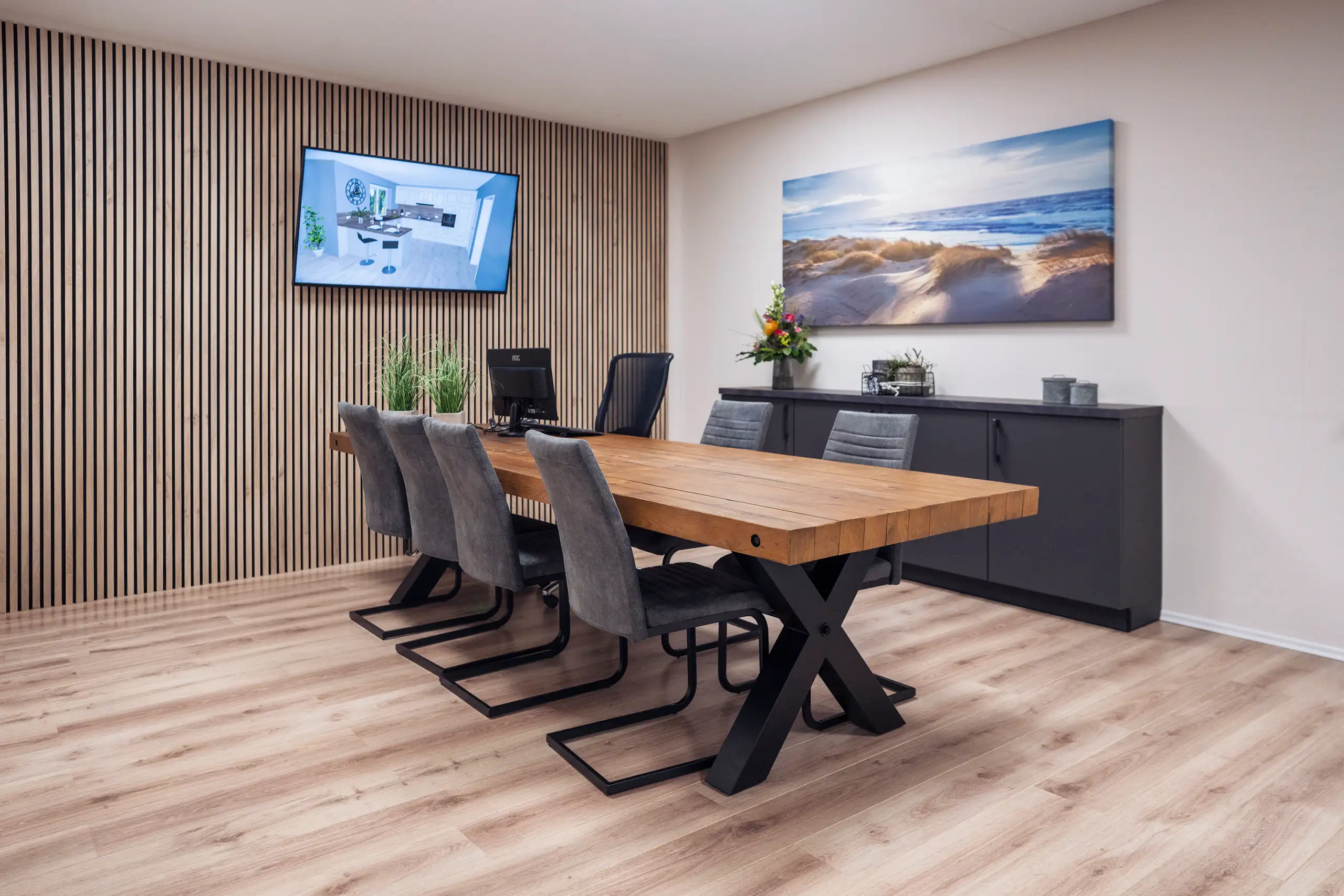 Moderner Konferenzraum mit großem Holztisch und sechs grauen Stühlen. An der Wand hängt ein großes Foto von einer Küste und ein Bildschirm. Der Raum hat einen Boden aus Holz und eine Wand mit Holzvertäfelung.